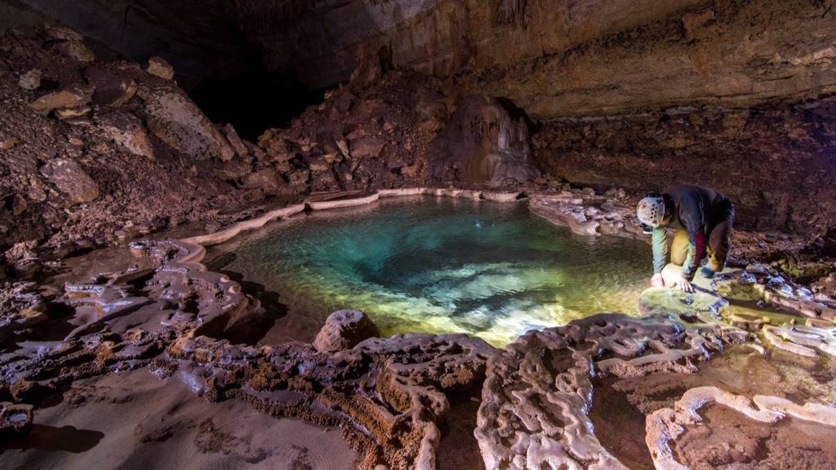 man in a helmet crouched next to a deep pool within a cave